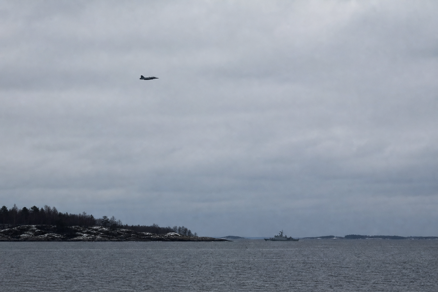 Harmaa merimaisema Suomenlahdella, taivaalla etäinen sotilaskoneen siluetti ja horisontissa viranomaisalus.
