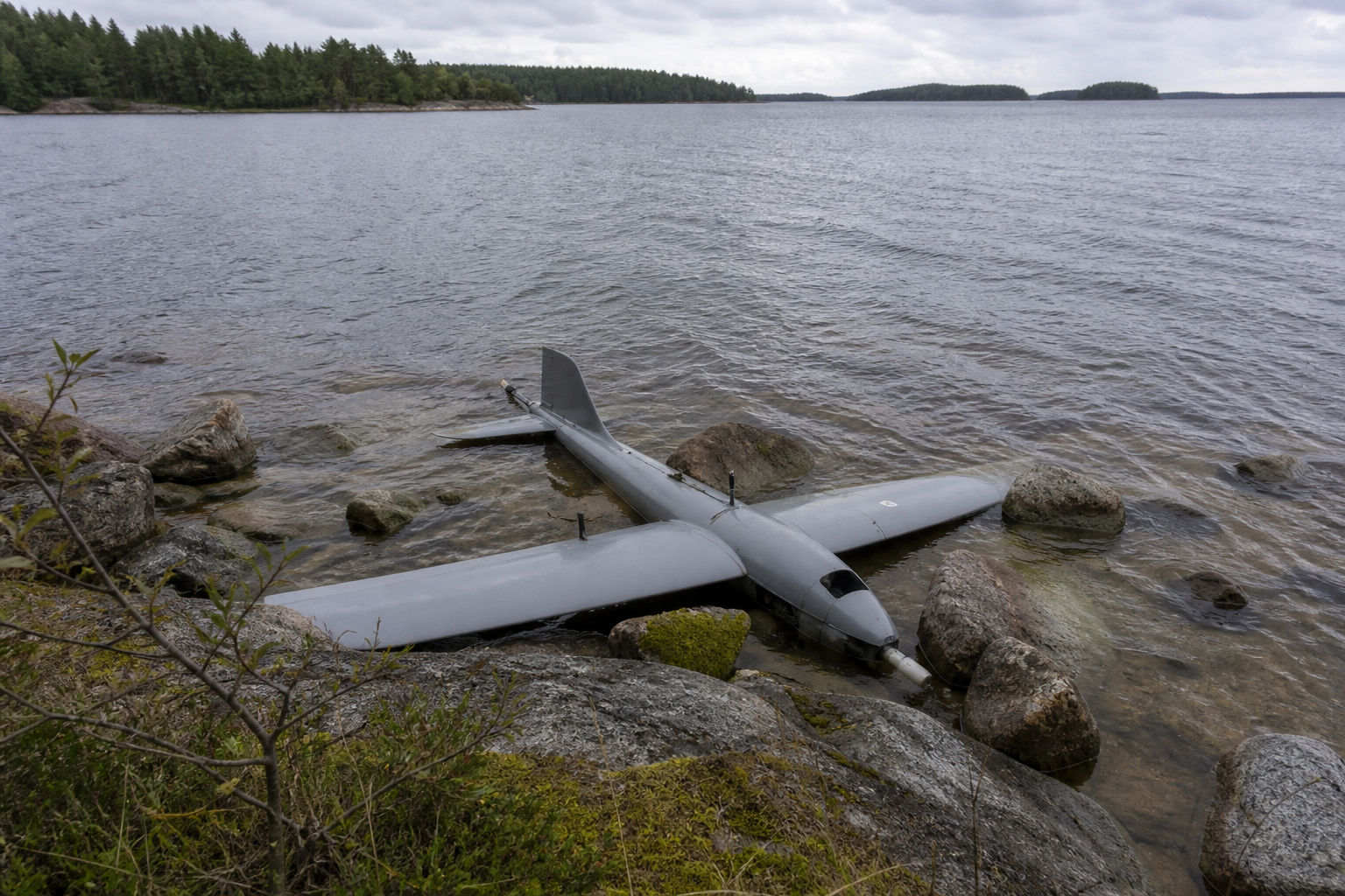 Tunnistamaton kiinteäsiipinen lennokki osittain kivikon ja kasvillisuuden seassa saariston rannalla, taustalla merta ja metsäisiä saaria.